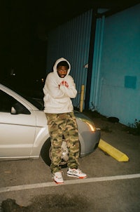 a man in a camouflage hoodie leaning against a car
